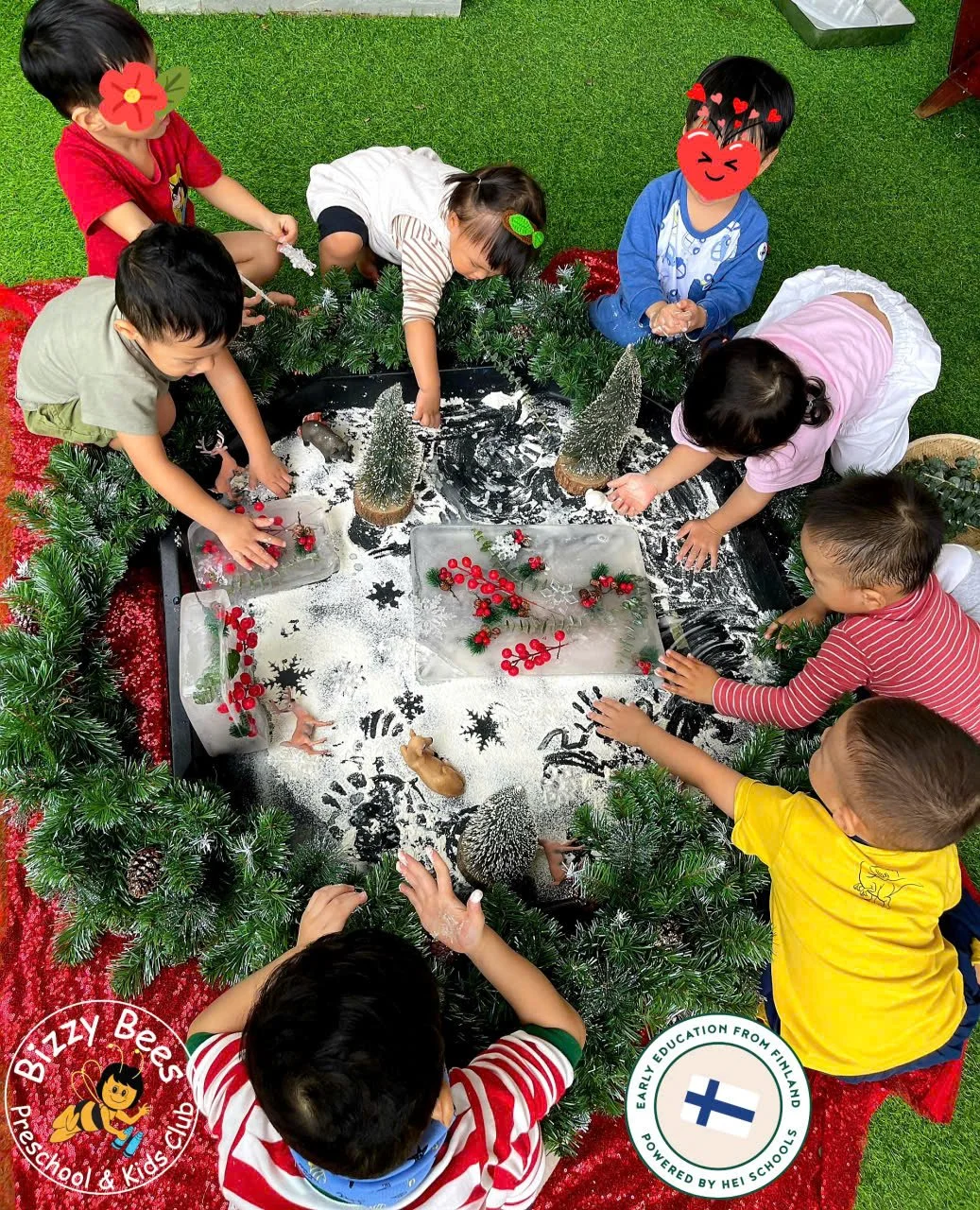 Children gathered around a small indoor winter scene with miniature Christmas trees, snow, berries, and animal figurines, creating a festive holiday display.