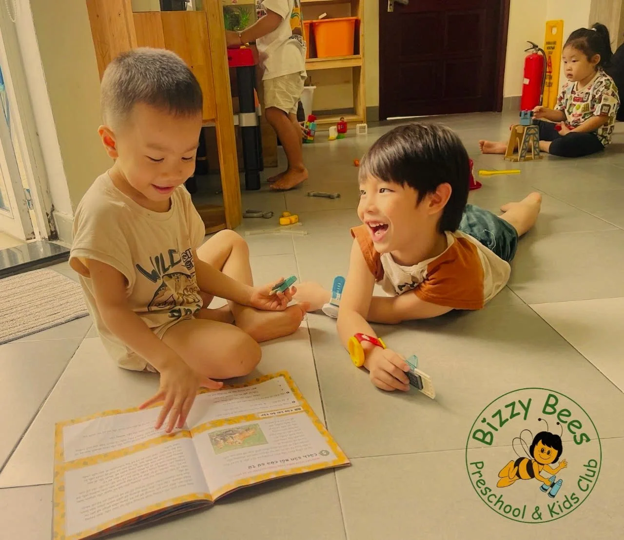 Two children smiling and engaging with a book and small toys. Other children and toys visible in the background