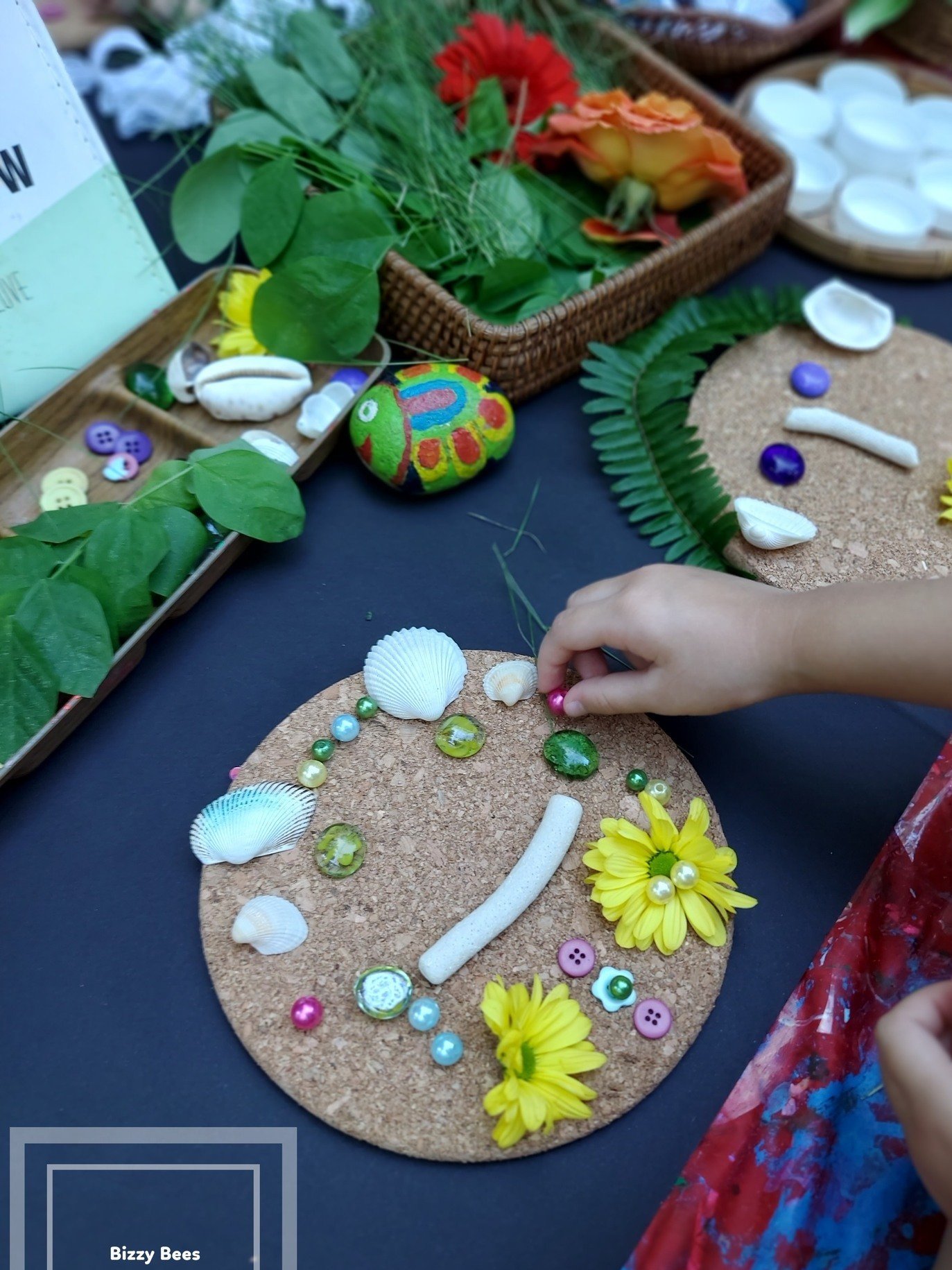Child decorating a circular corkboard with seashells, beads, and flowers at a craft table.