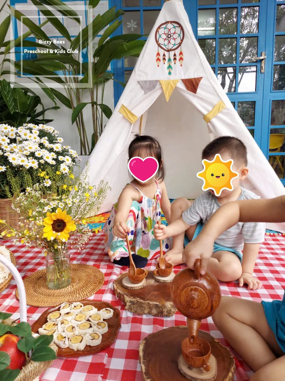 Two children playing with toy wooden tea set inside a white teepee tent decorated with a dreamcatcher, with a checkered tablecloth, flowers, and snacks on a picnic setup.