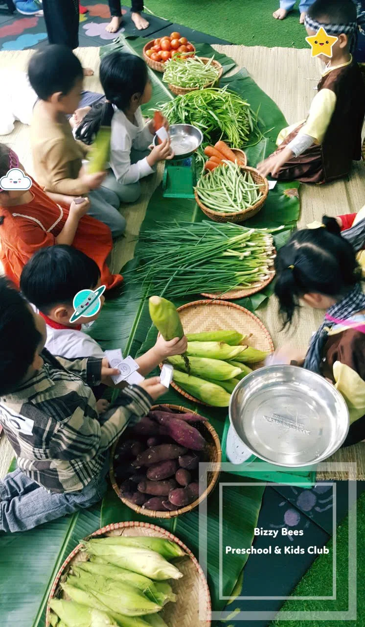 Children sitting on a mat around a table of fresh vegetables including corn, carrots, green beans, sweet potatoes, and tomatoes, at Bizzy Bees Preschool & Kids Club.