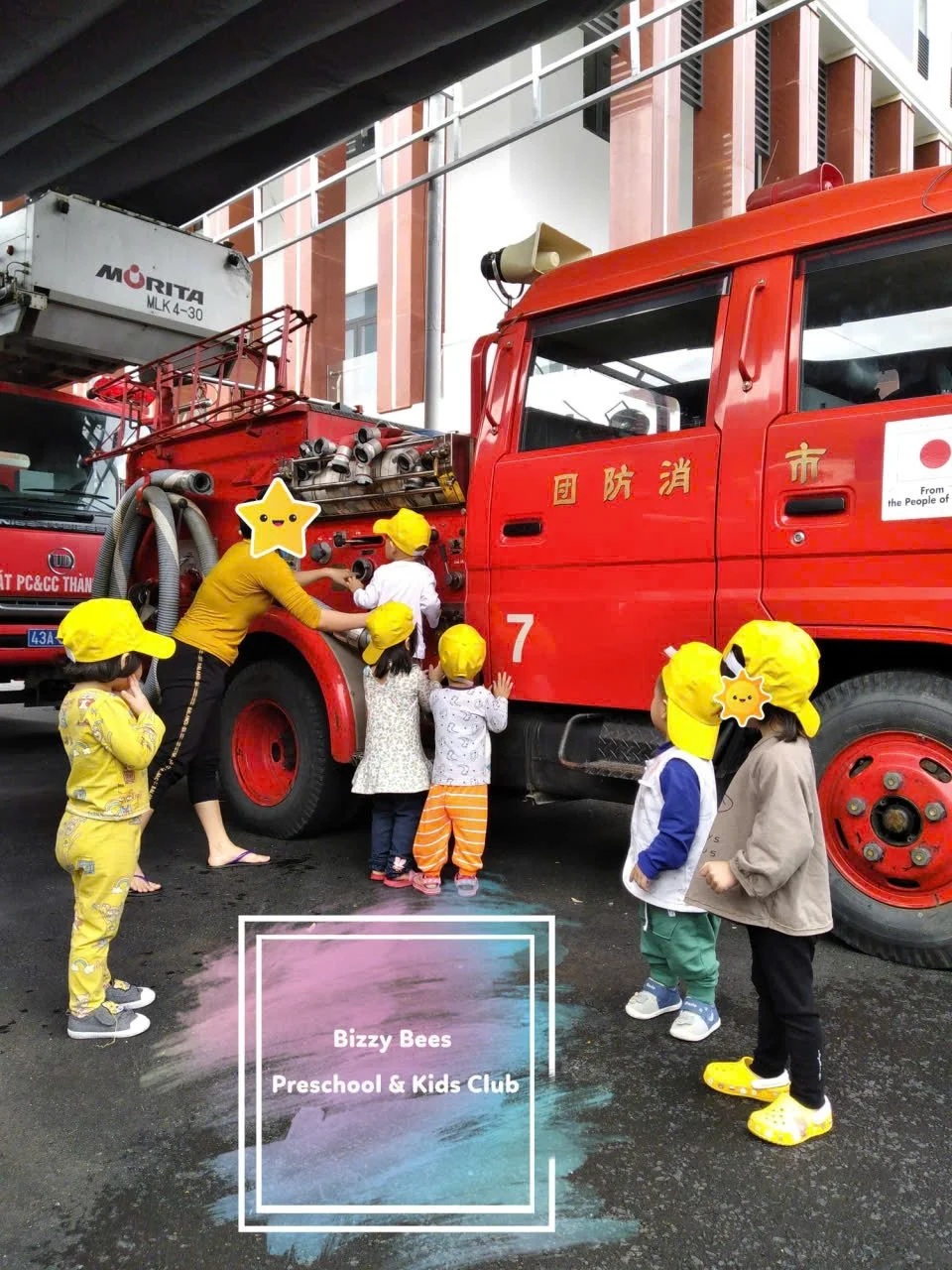 A group of children wearing yellow hats gathered around a red fire truck, with an adult assisting them. The scene is outdoors in an urban area.