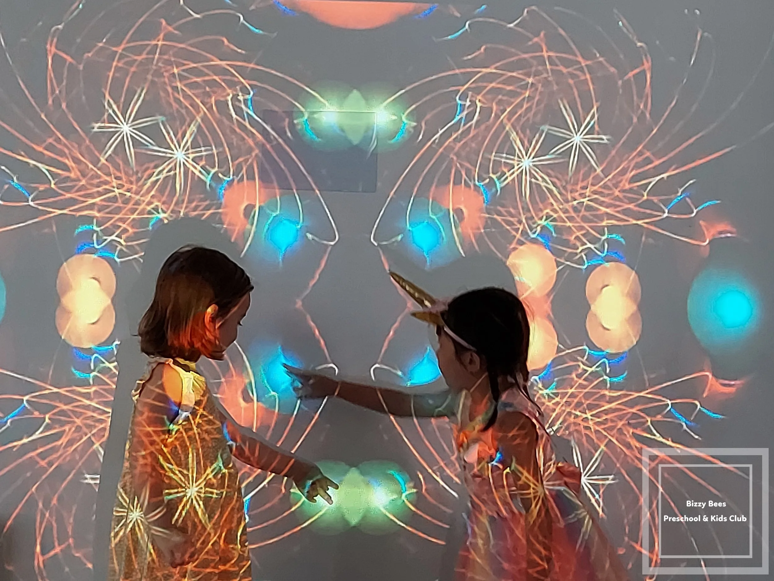 Two young children are playing in front of a colorful, abstract light projection that resembles fireworks. 