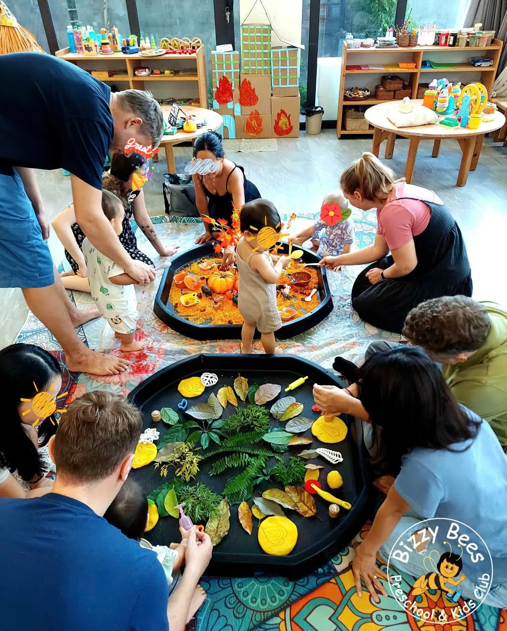 Children and adults gathered around two sensory play tables with autumn and nature-themed items, in a classroom or playroom setting.