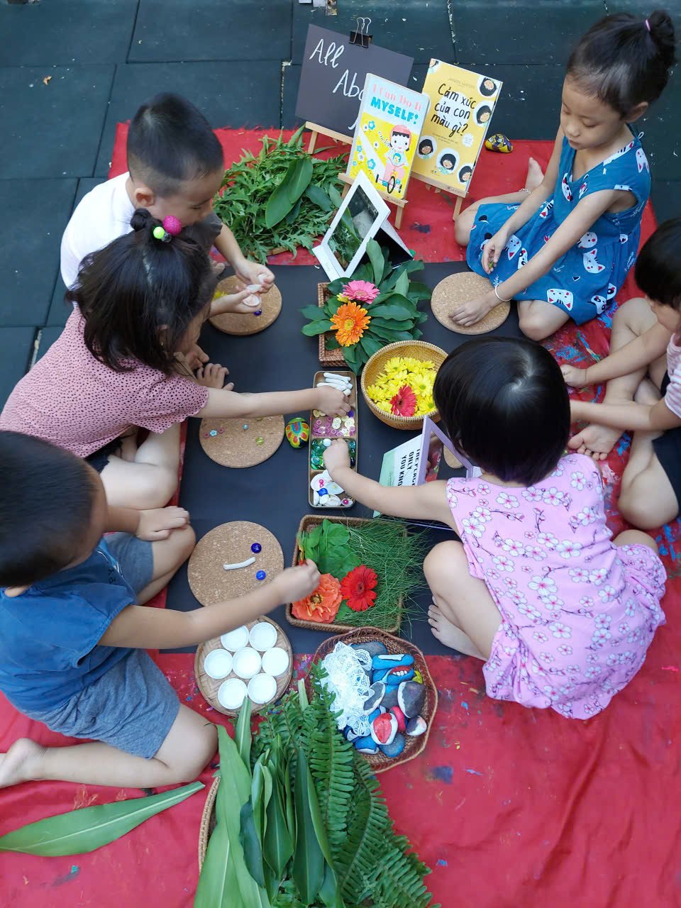 Group of children sitting on a red cloth and engaging in arts and crafts, surrounded by flowers, leaves, and craft supplies, with various signs and pictures displayed in the background.