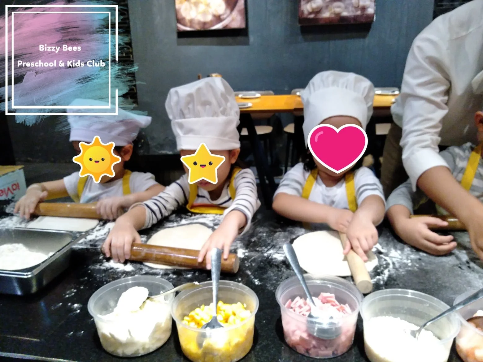 Children wearing chef hats and aprons, sitting at a table, rolling out pizza doughs