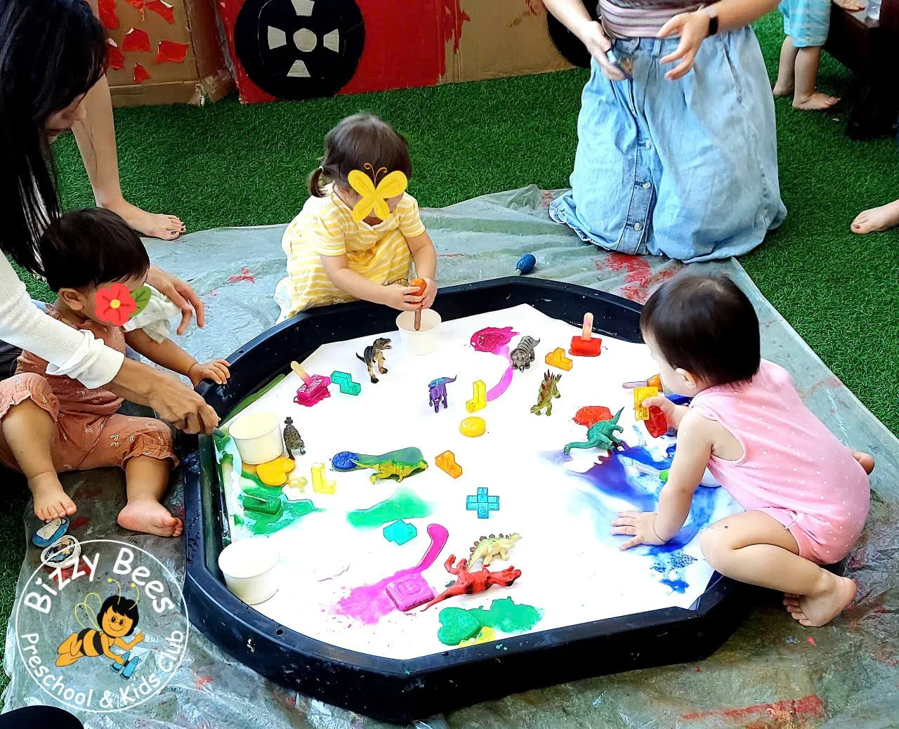 Children playing with colorful toy dinosaurs and paint on a large white tray outdoors. An adult supervises, and a logo for 'Bizzly Bees Preschool & Kids Club' is visible in the corner.