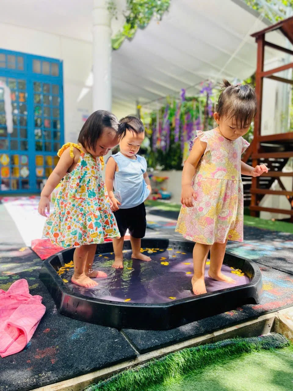 Three young children standing in a shallow water tray outdoors, playing and exploring.