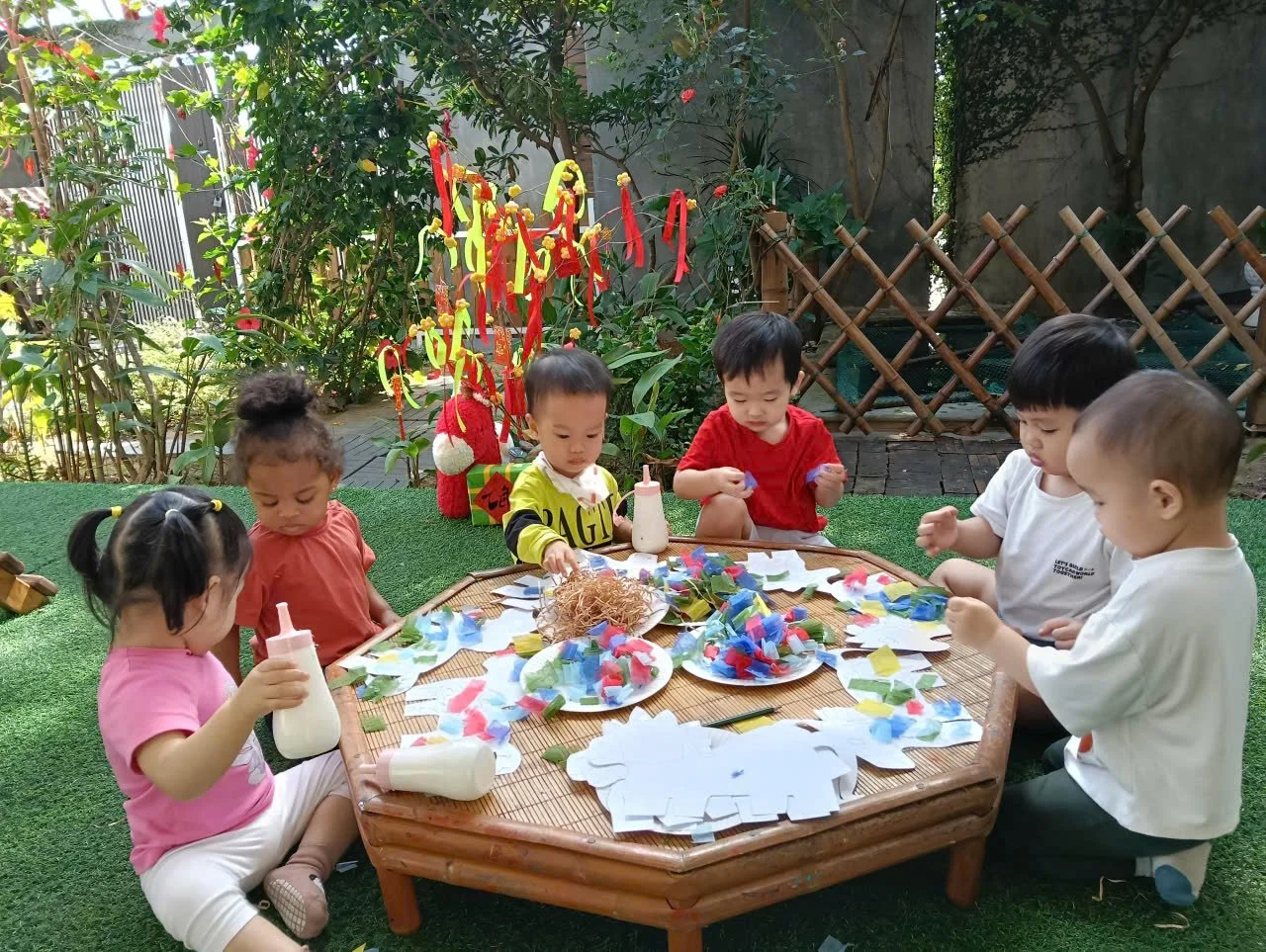 Six young children gathered around a round outdoor table, engaged in arts and crafts with colorful paper pieces and glue bottles, in a garden setting with greenery and decorative hanging ribbons.