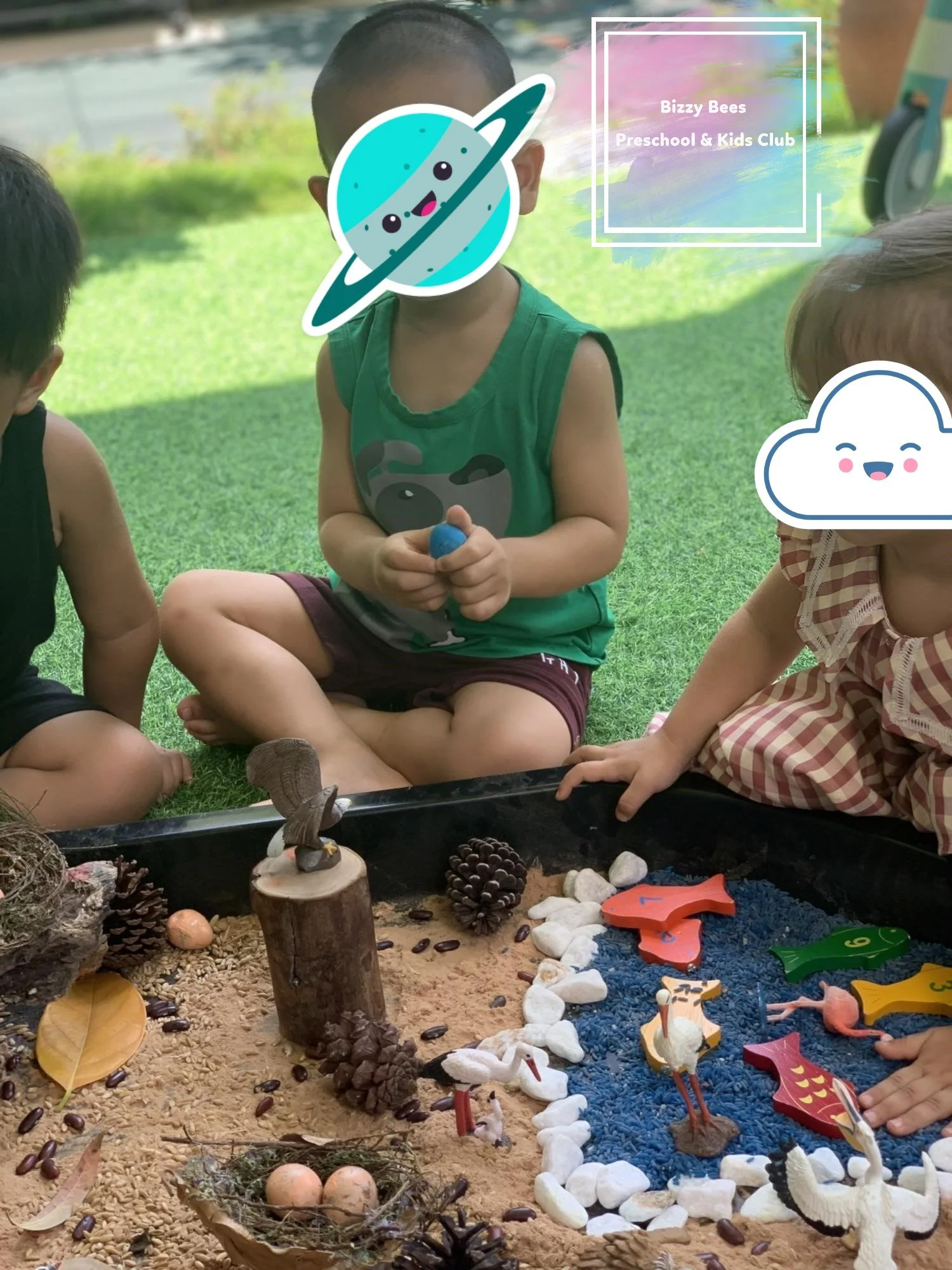Children playing with a sandbox filled with small toys, pinecones, rocks, and decorative items, outdoors on green grass.