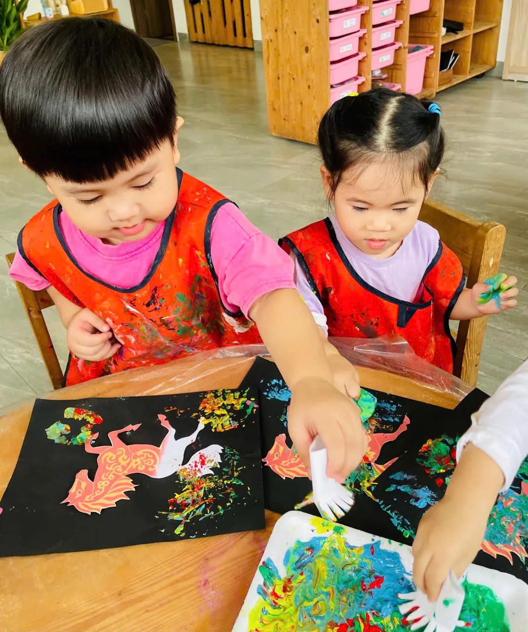 Two young children, a boy and a girl, engaging in a painting activity at a table in a classroom. They are wearing red smocks and focused on creating colorful art on black paper using paint and sponges.