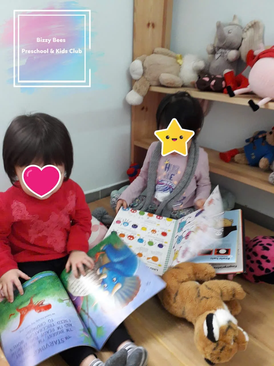 Two young children sitting on the floor in a playroom, reading a colorful picture book surrounded by plush toys, with a shelf of stuffed animals behind them. The image features stickers covering the children's faces for privacy.