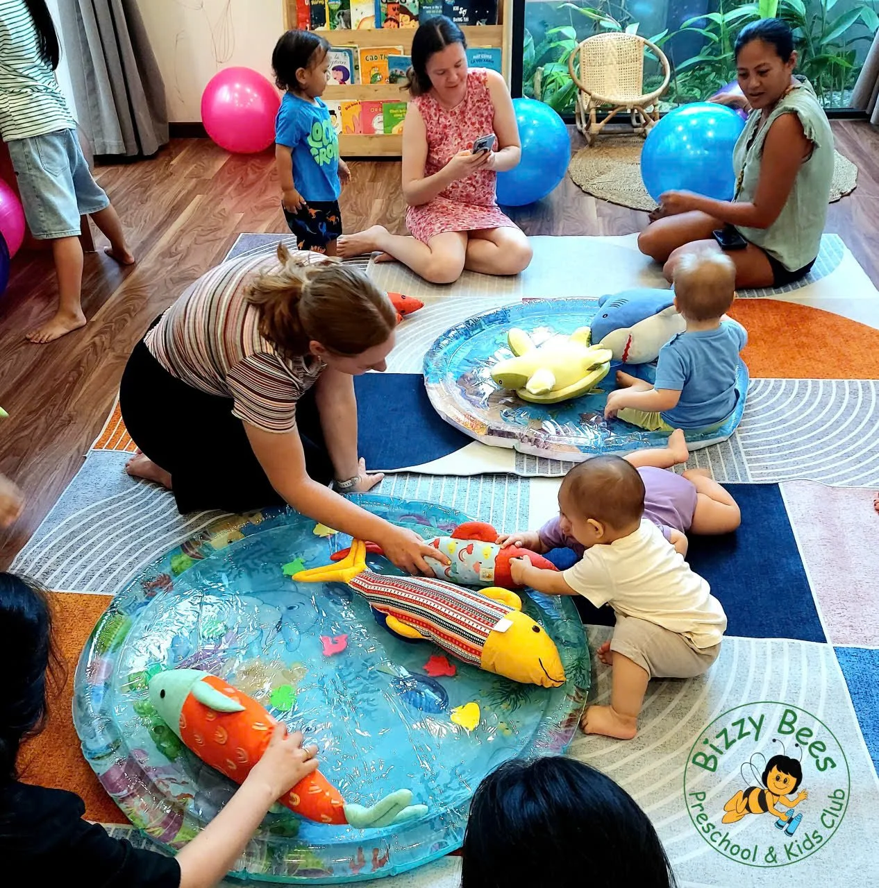 Children and adults gathered around small inflatable pools with plush fish toys, indoors with a bookshelf, large green plants, and pink and blue exercise balls in the background. Every person appears engaged in play or observing.