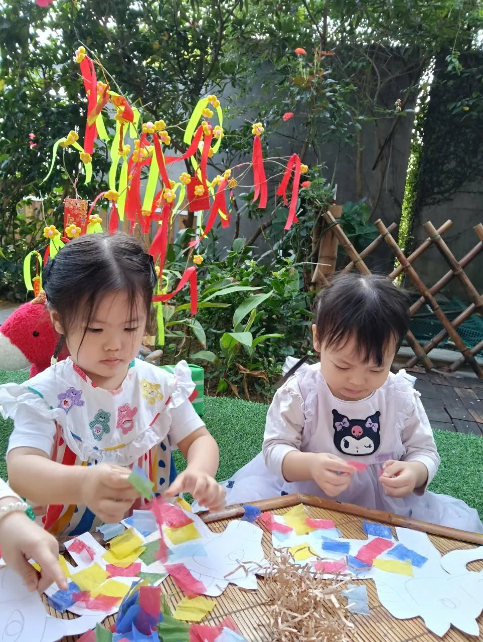 Two children sitting at a basket, decorating paper cutouts with colorful tissue paper, in a garden with hanging ornaments and greenery in the background.