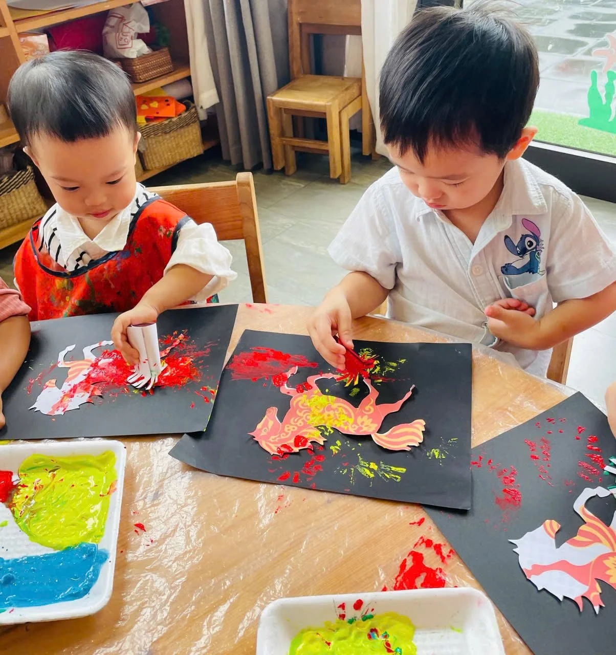 Two young children engaging in an art activity, coloring paper with red, yellow, and green paints, sitting at a wooden table in a classroom or art room.