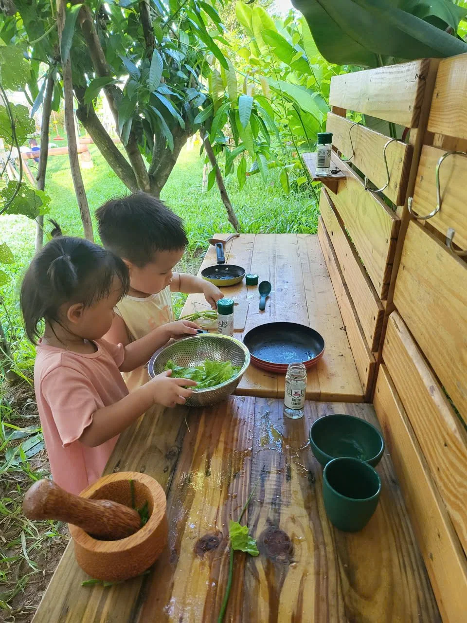 Two young children, a girl and a boy, preparing a salad outdoors at a wooden table surrounded by lush green foliage. The girl is holding a colander filled with leafy greens, and the boy is reaching for a spice jar. The table has bowls, a mortar and p