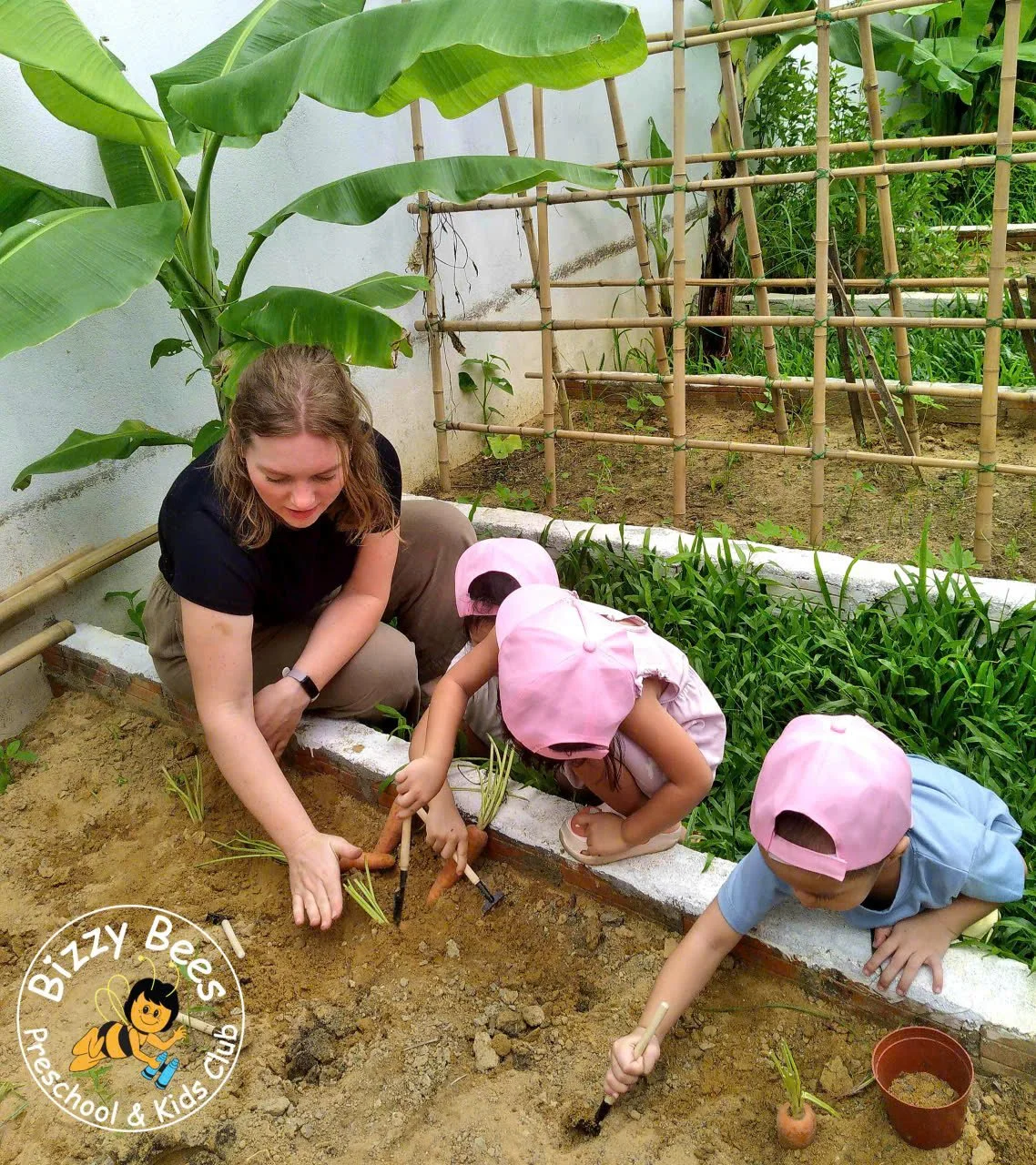 A woman and two young children planting carrots in a garden bed. The children are wearing pink hats and the woman is guiding them as they insert the vegetables into the soil. The garden has green plants, banana leaves, and bamboo fencing in the backg