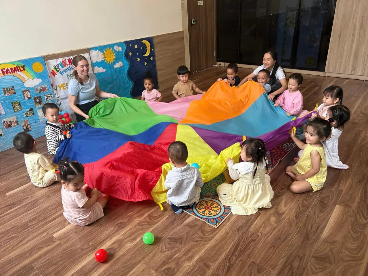 Children and teachers playing with a colorful parachute in a classroom.