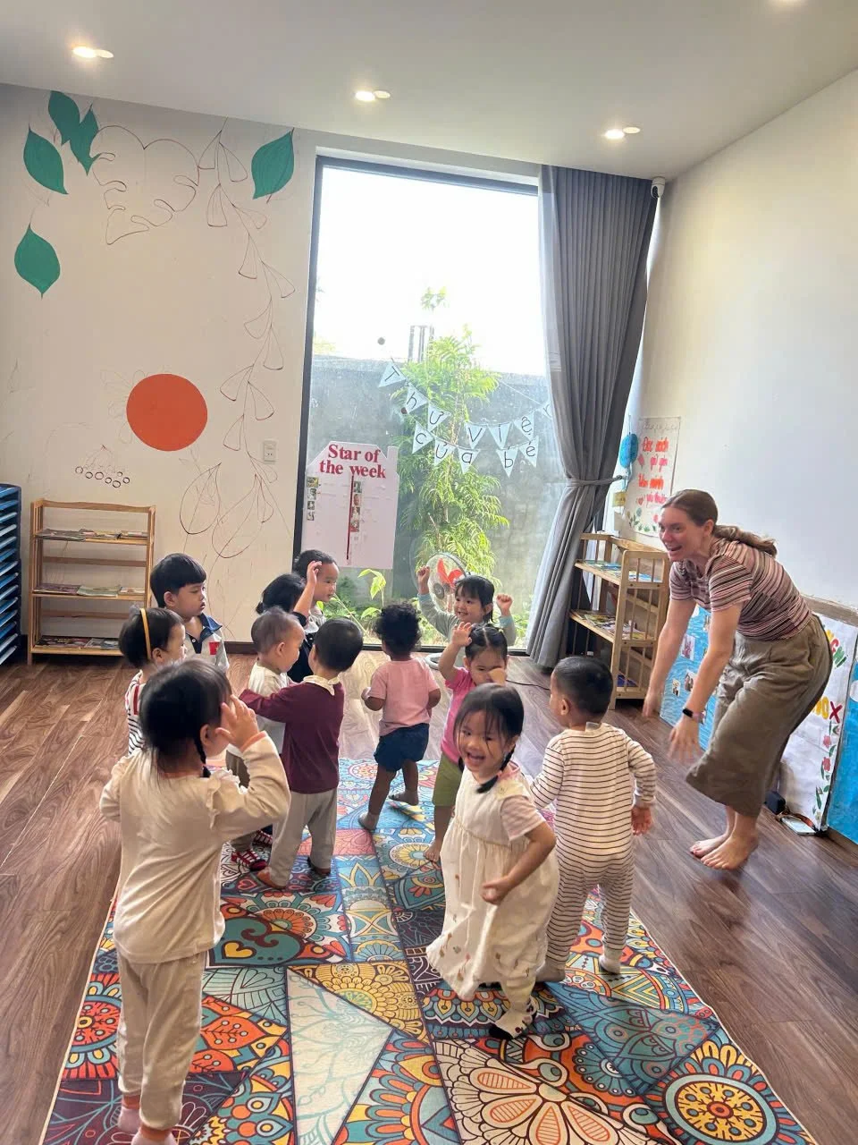 A group of young children dancing and playing in a classroom with a colorful patterned rug, a teacher participating, and decorations on the walls including a 'Star of the Week' poster and a window with blue curtains.