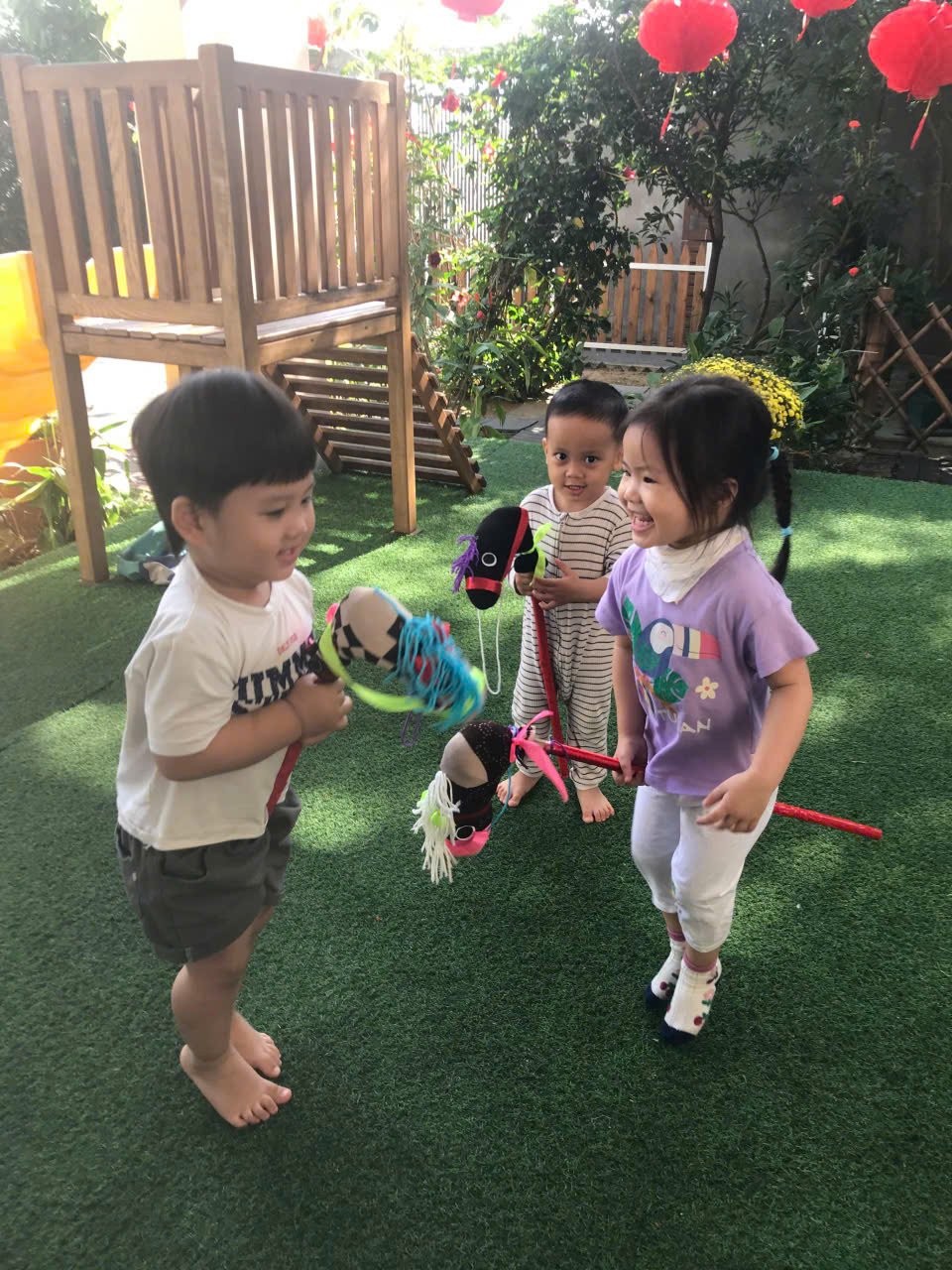 Three young children playing with sock puppets outdoors on a grassy yard, surrounded by plants and trees, with a small wooden play structure in the background.