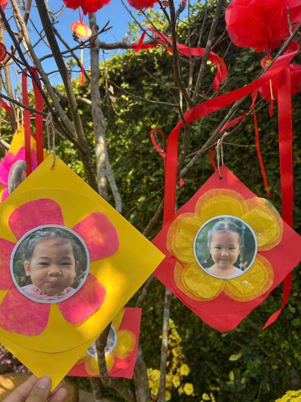 Colorful paper lanterns with children's faces inside flower-shaped cutouts, hanging from tree branches for a celebration or festival.