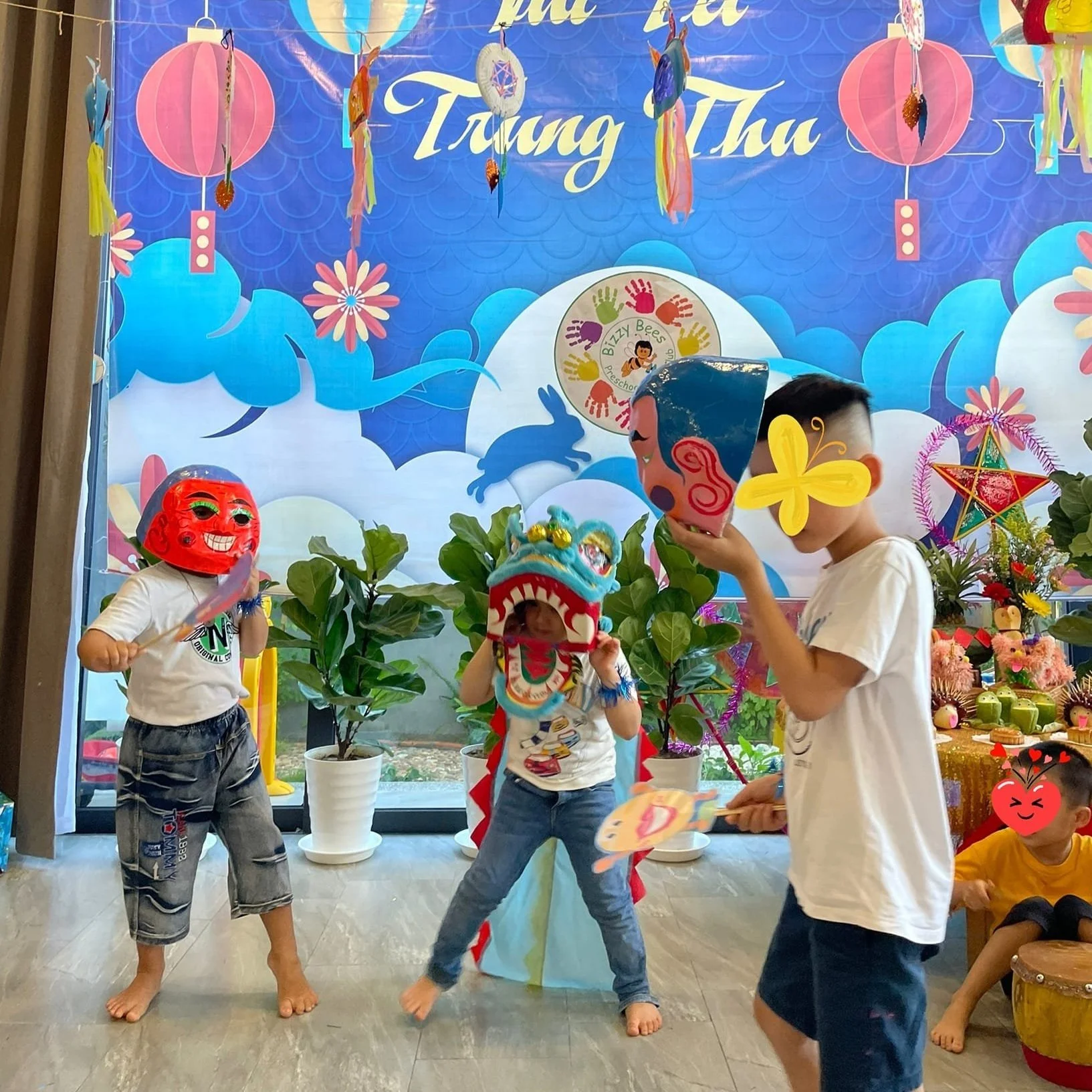 Children wearing colorful masks and costumes participate in a dragon dance for the TET festive celebration, with vibrant decorations including lanterns and stars in the background at Bizzy Bees Pre-school and kids club in Da Nang