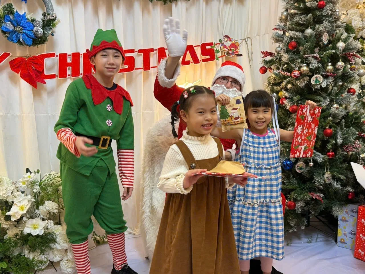 Children dressed in Christmas costumes standing with Santa Claus in front of a decorated Christmas tree, holding gifts and presents, smiling during a holiday celebration.