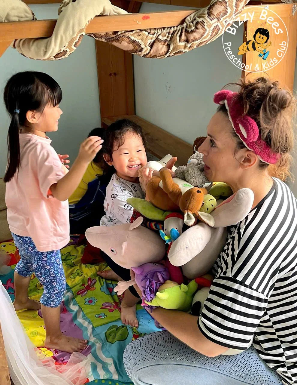 A woman and two young girls playing with stuffed animals on a colorful bed in a playroom. The woman is wearing a striped shirt with a pink headband and has a playful expression. One girl is smiling and the other girl is standing, smiling, and reachin