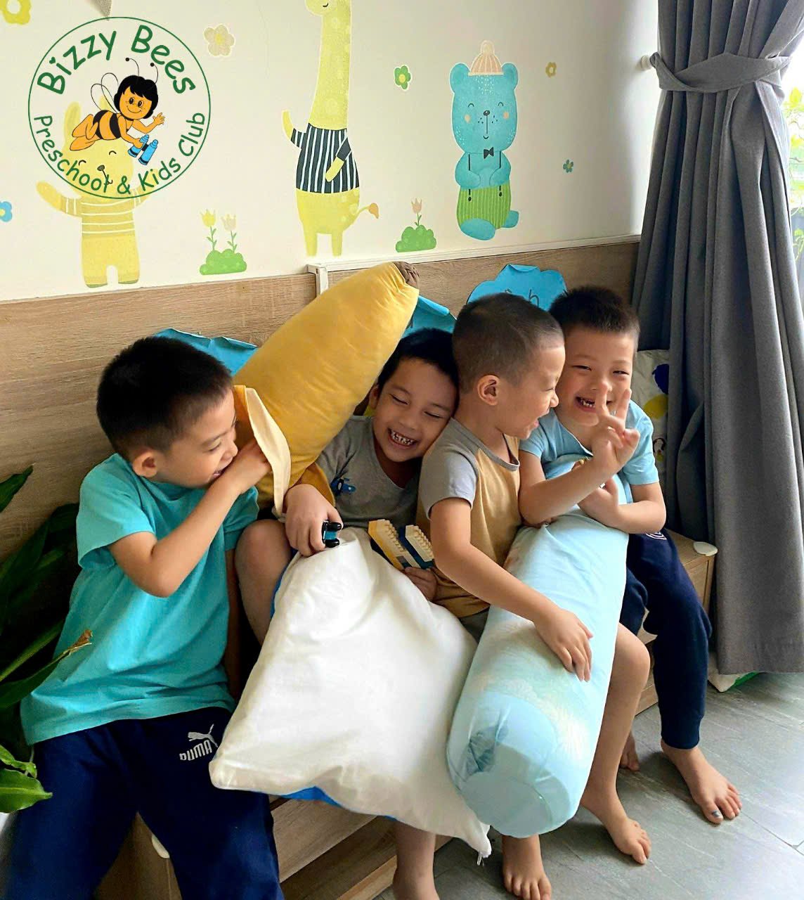 Four young boys sitting on a bed, laughing and playing with pillows. The wall behind them is decorated with colorful animal and tree illustrations, and there is a curtain on the side. The banner reads 'Bizzy Bees Preschool & Kids Club.'