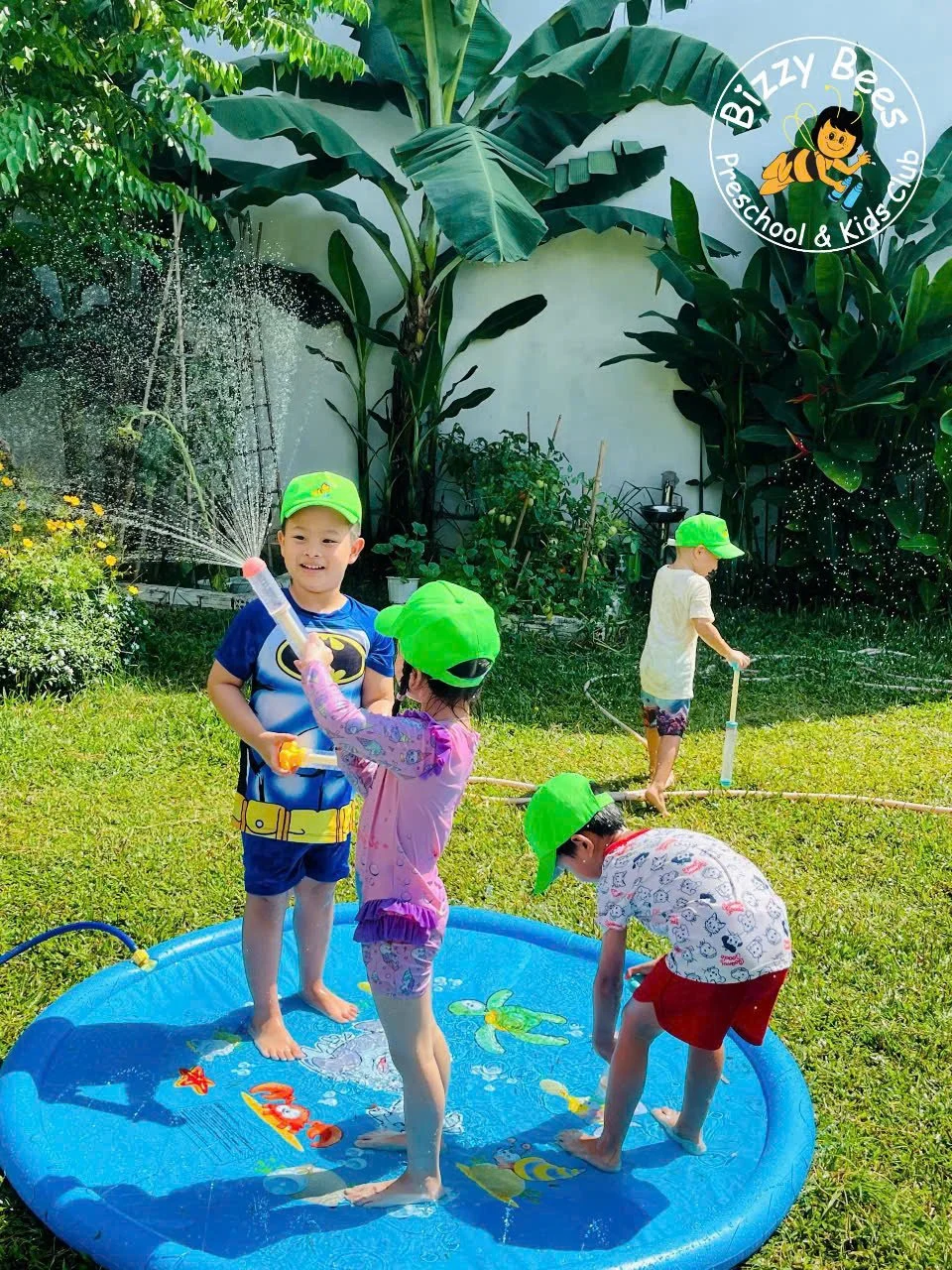 Four children playing with water in a small blue plastic pool in a backyard garden, wearing green hats. Two children are standing in the pool, one boy and one girl, with the girl spraying water toward the boy. Another boy is outside the pool, leaning