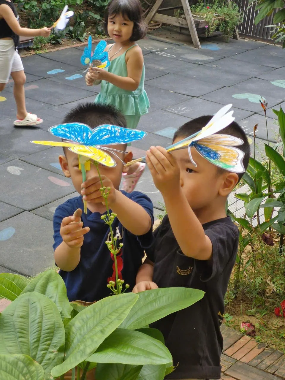 Two children holding painted paper butterfly cutouts in front of their faces, with a girl standing in the background holding a similar butterfly. They are outdoors near green plants and large leaves, on a tiled surface with colorful chalk markings.