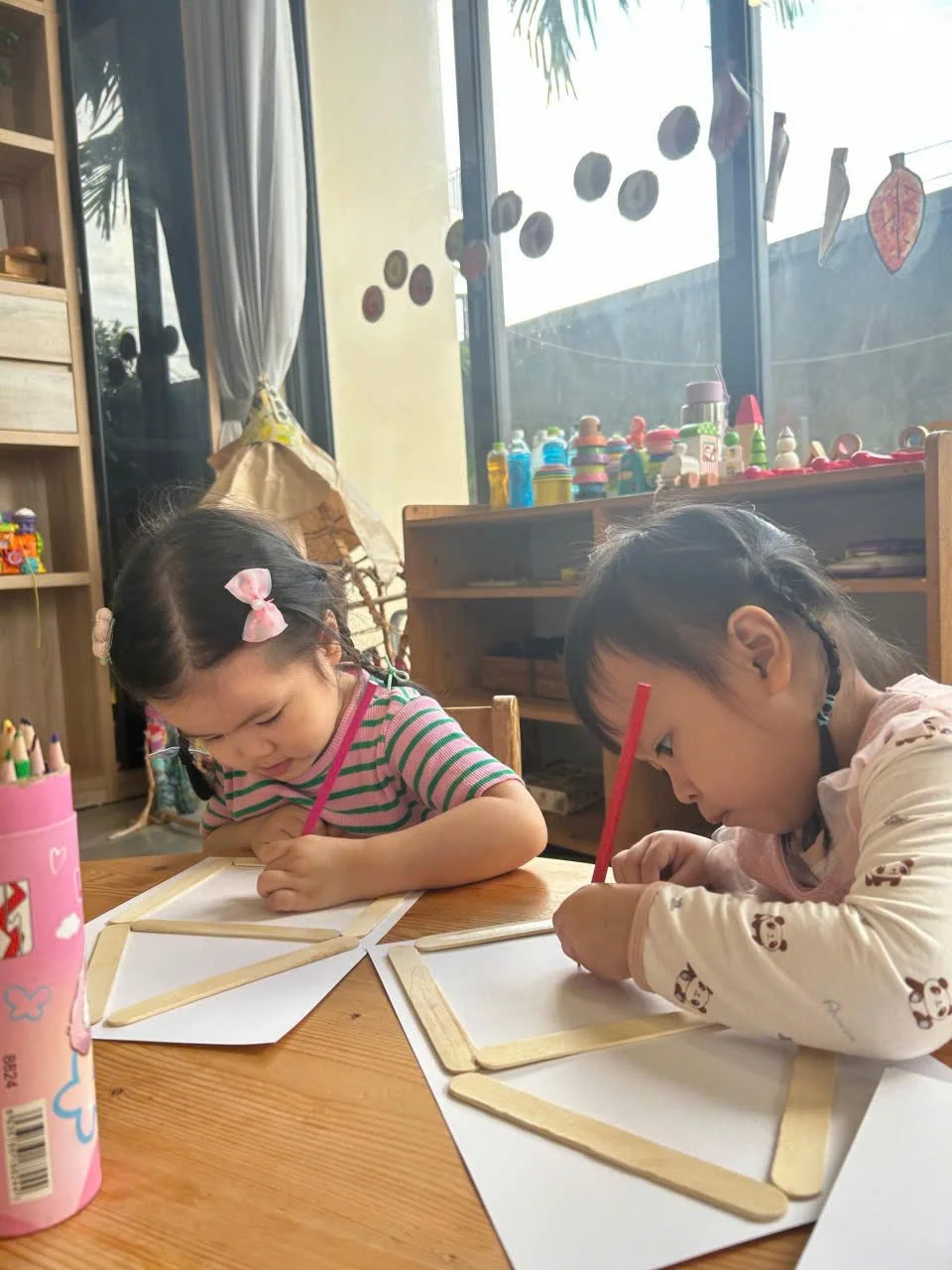Two young girls sitting at a wooden table, drawing on white paper with red pencils in a classroom. Behind them, large windows reveal a sunny outdoor scene with plants and toys on a shelf.