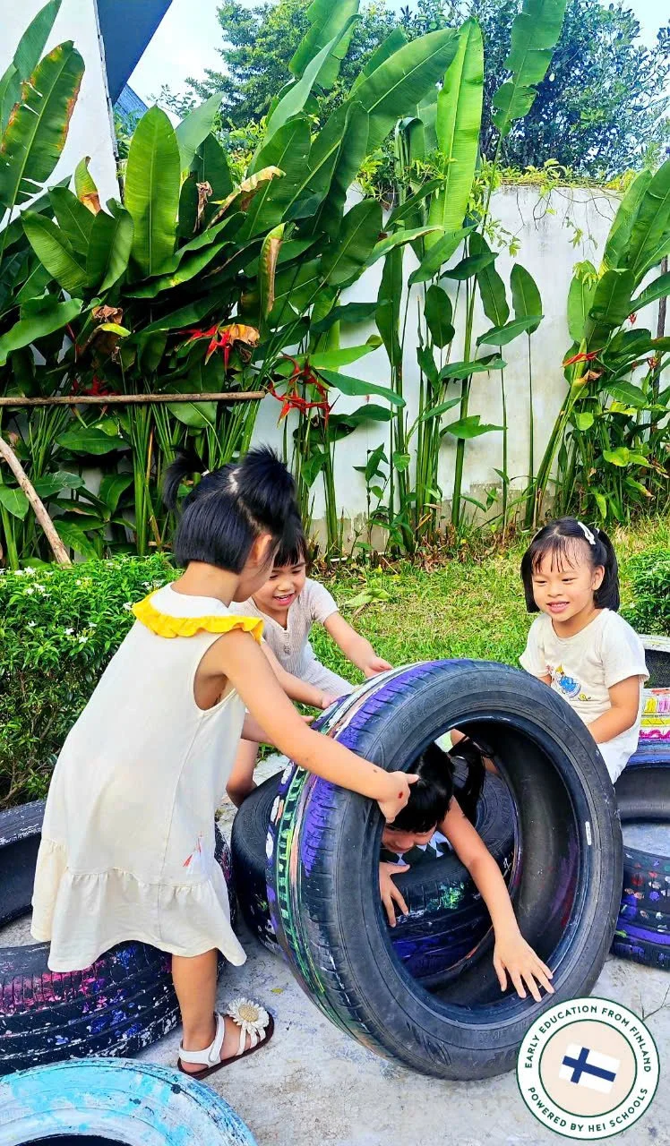 Four young children playing with recycled tires outdoors, surrounded by lush green plants at Bizzy Bees KIndergarten