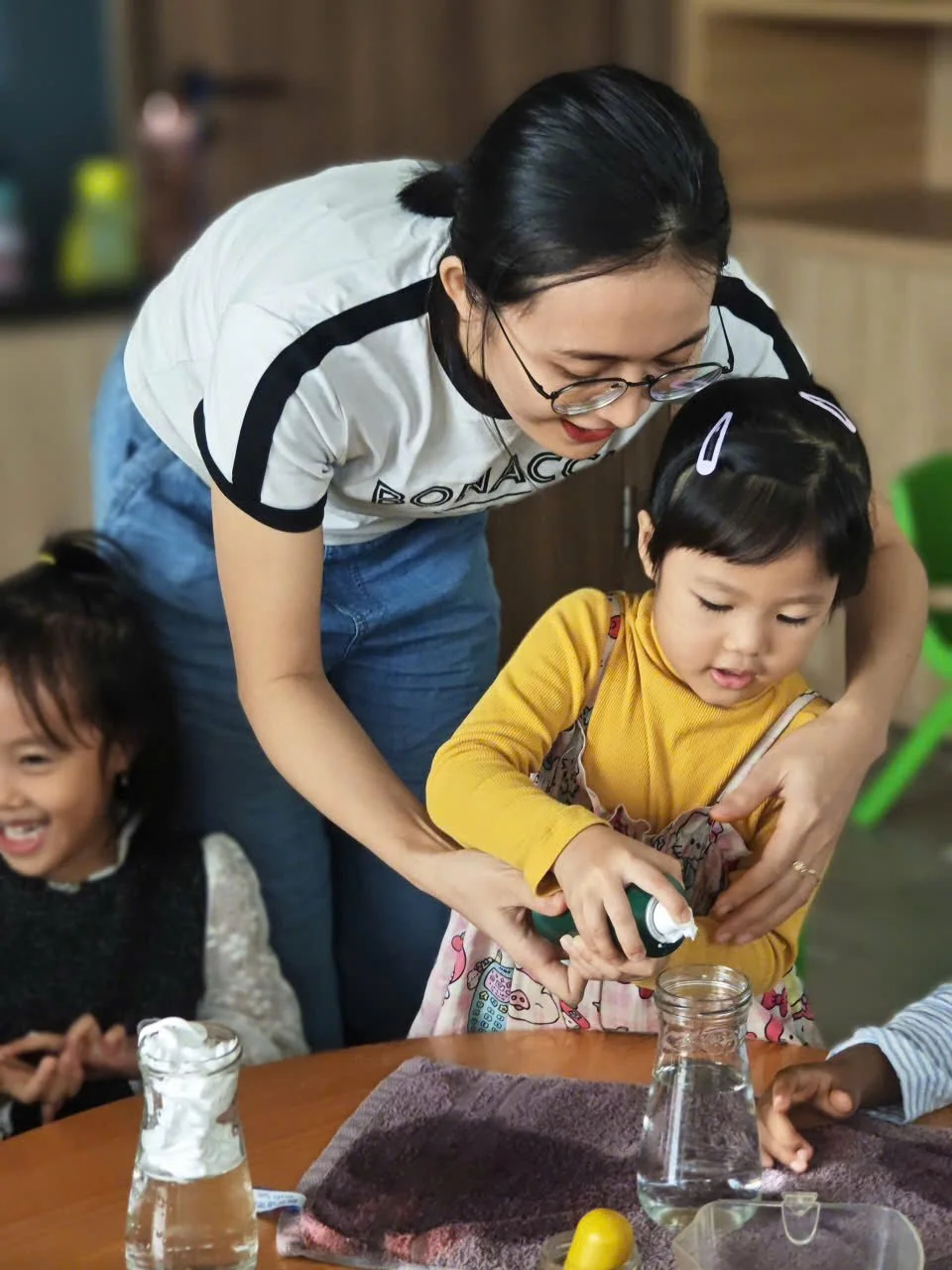 A woman assisting a young girl with a science experiment at a table, with two other children nearby, in a classroom or learning environment.