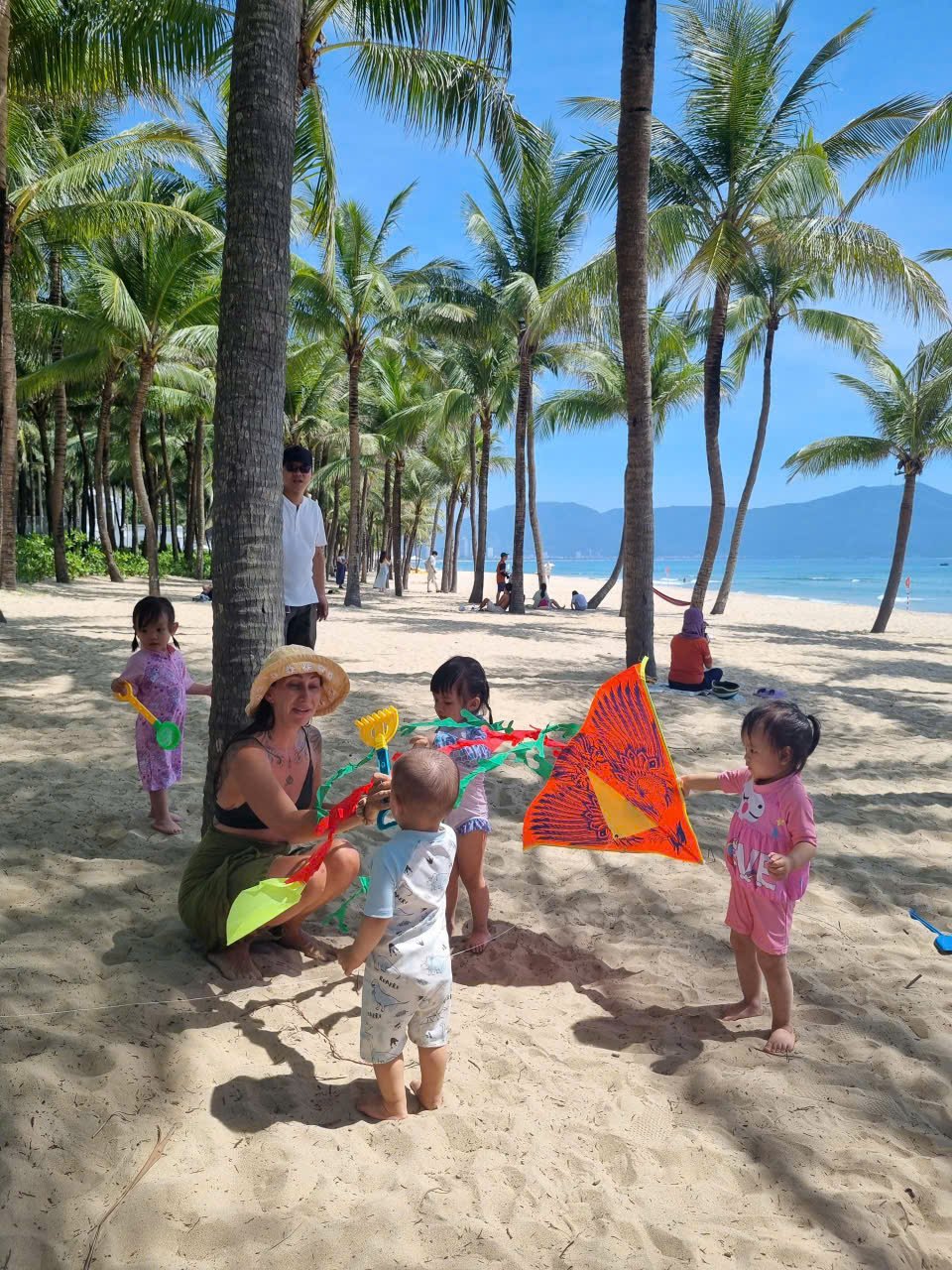 Children and adults playing with a colorful kite on a sandy beach with palm trees, blue sky, and ocean in the background.