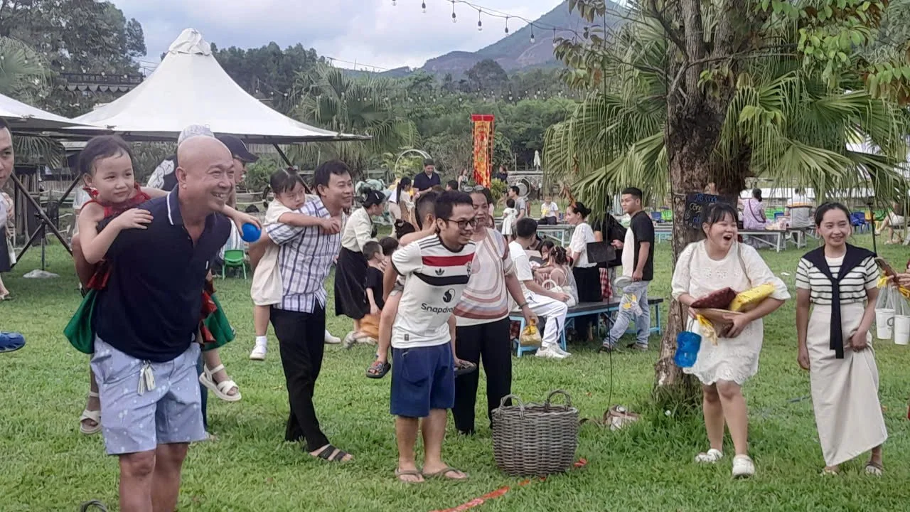 Group of people enjoying a festive outdoor gathering under a tree, with some carrying children and others holding food and drinks, in a lush, green setting with tents and string lights overhead.