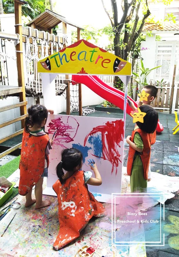 Four young children engaged in painting on a large whiteboard outdoors, with a colorful 