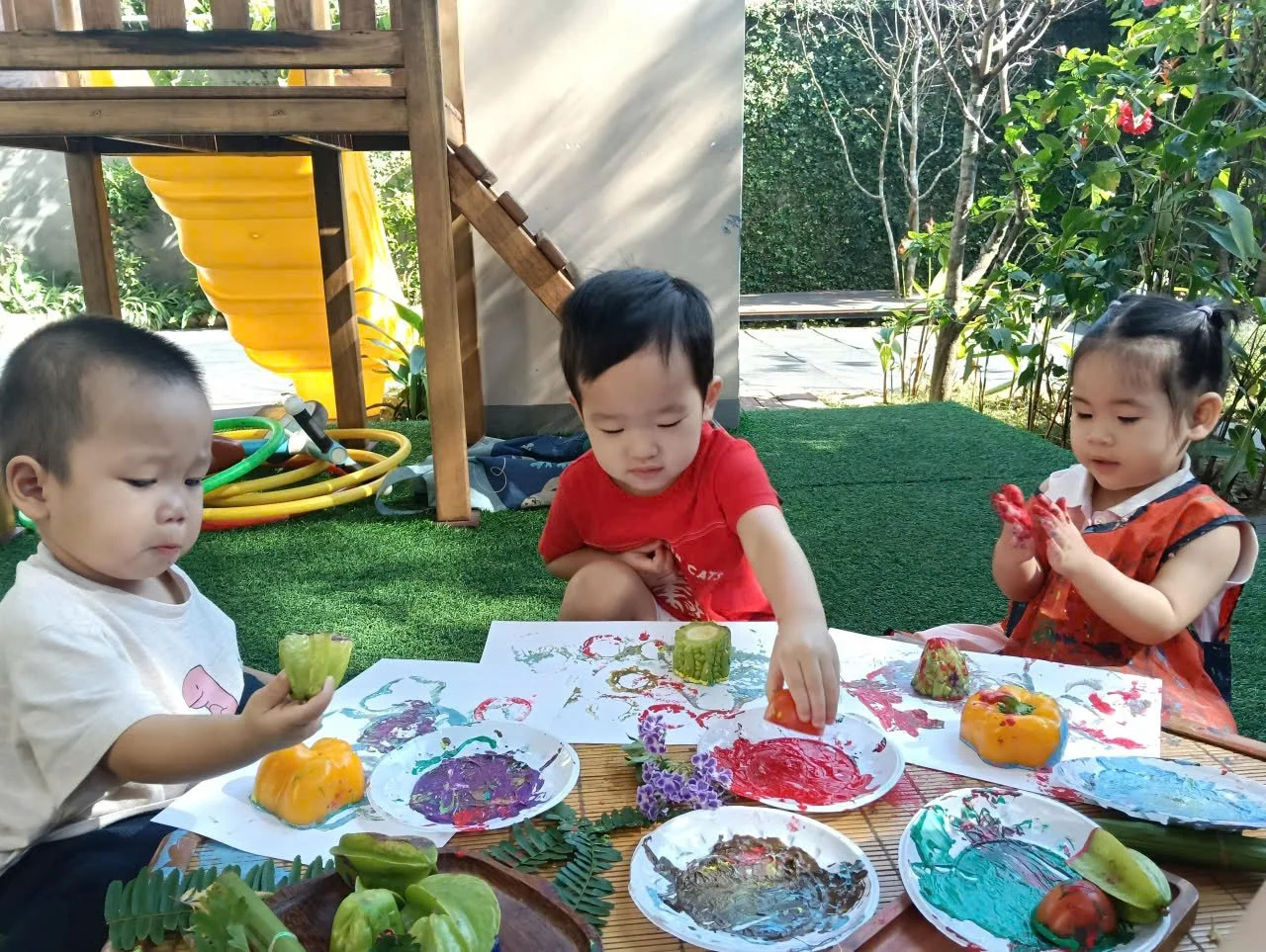 Three young children painting and decorating vegetables outdoors on a table with plates of colorful paints, surrounded by greenery and play equipment.
