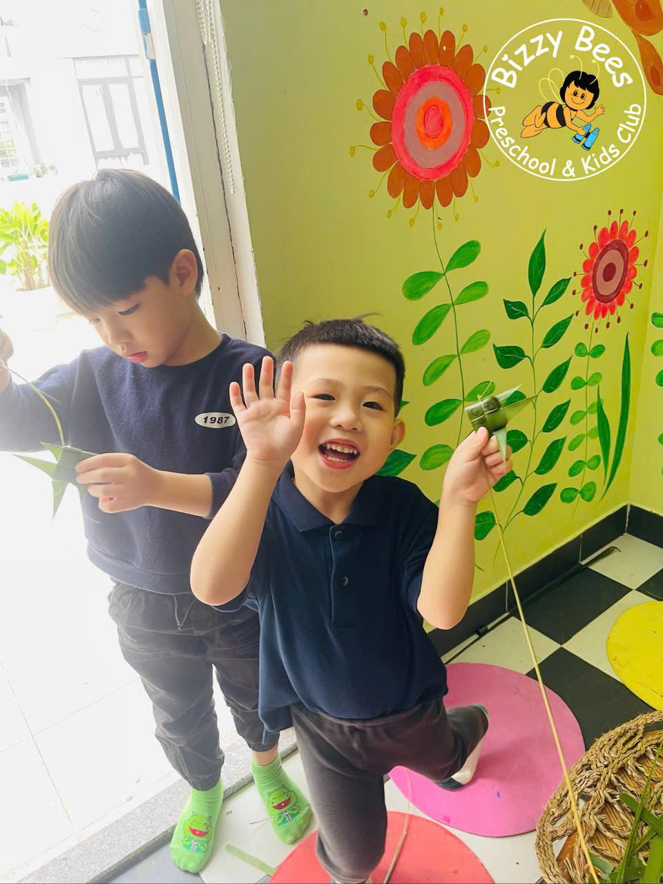Two young boys smiling, one waving, holding a kite inside a colorful preschool classroom with floral wall decorations.