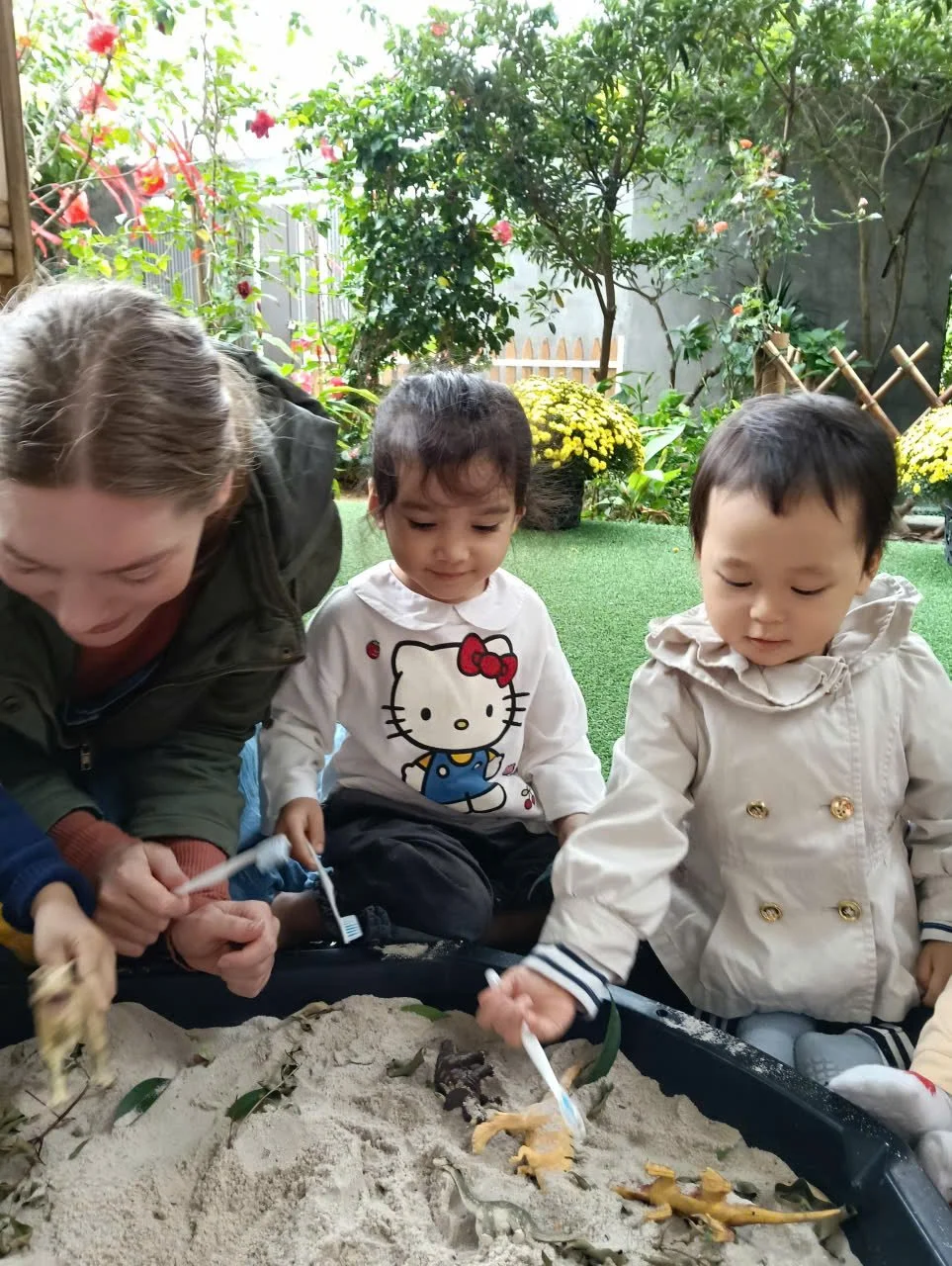 Children and a teacher playing with toy dinosaurs in a sandbox outdoors surrounded by plants and flowers.