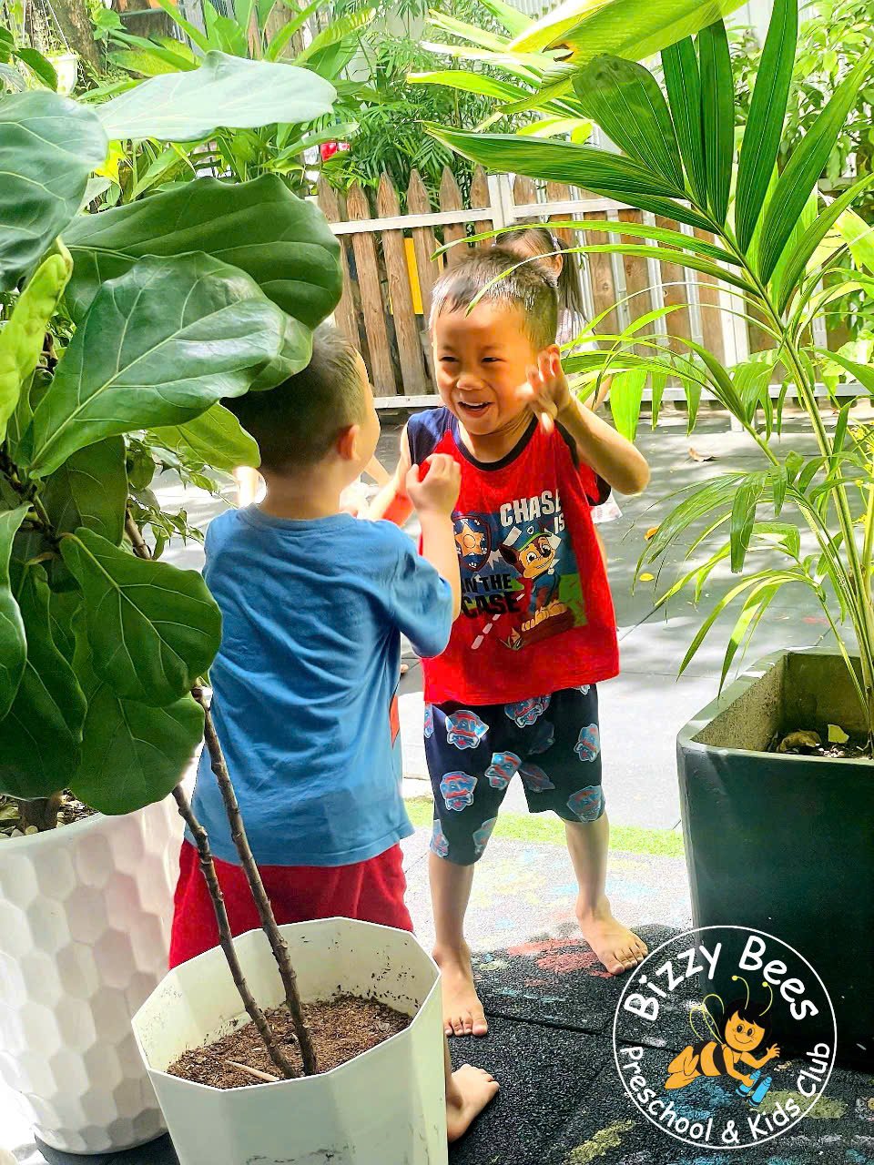Two young boys playing and laughing outdoors near potted plants, with a wooden fence in the background. One boy in a red shirt and shorts, and the other in a blue shirt and red shorts, appear to be having fun.