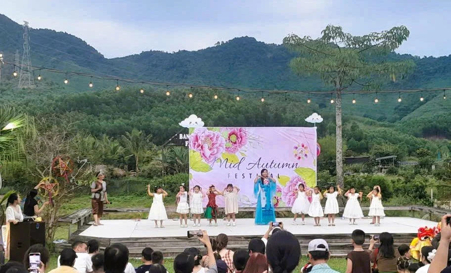 Outdoor stage set against green mountains during Mid Autumn Festival with children in costumes performing and audience watching.
