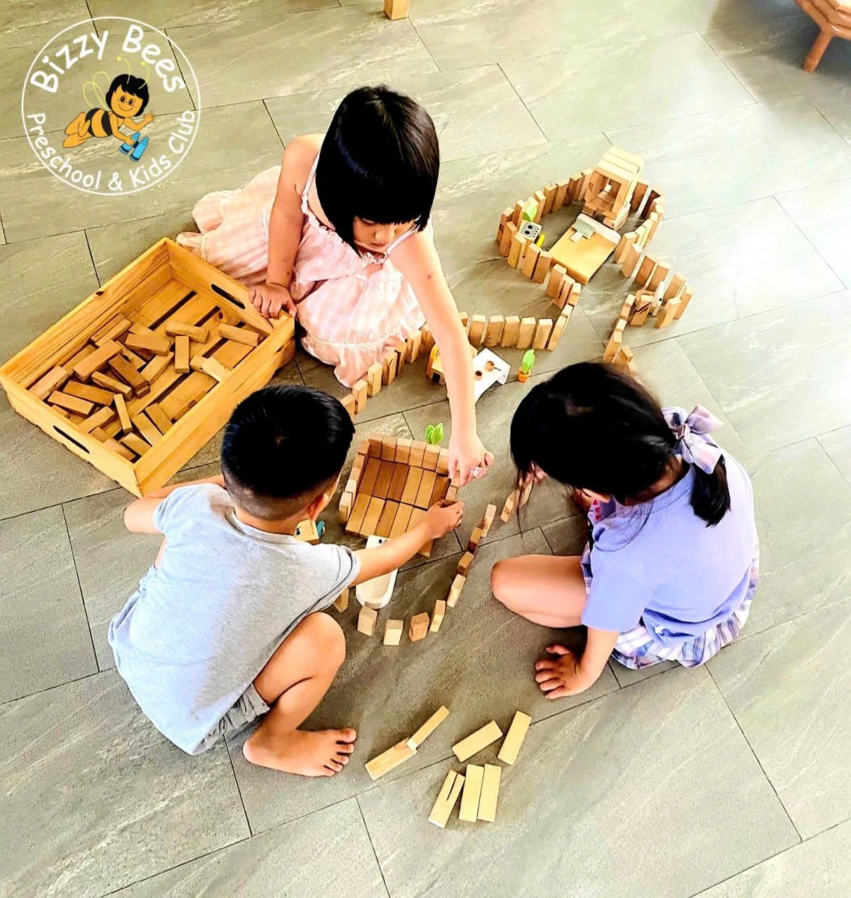 Three children playing with wooden blocks on the floor, building a structure. One girl, wearing a pink dress, is reaching out to place a block. A boy in a gray shirt and shorts, and another girl in a lavender outfit with a hair bow are also involved 