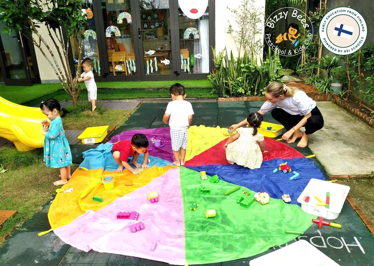 Children playing on a colorful parachute outside a preschool, with a teacher assisting. There are toys on the parachute, a yellow slide to the left, and a garden with trees and plants in the background.