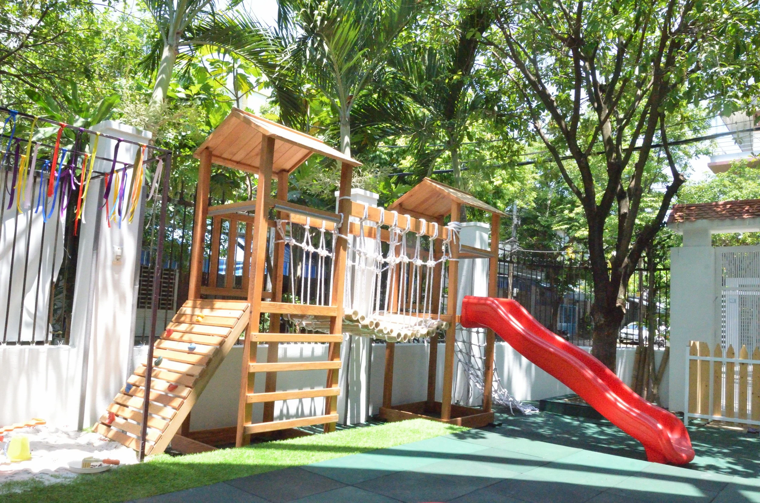 Children's outdoor playset with slide, ropes, climbing wall, and shaded areas, set in a backyard with white fence, lush green trees, and morning sunlight.