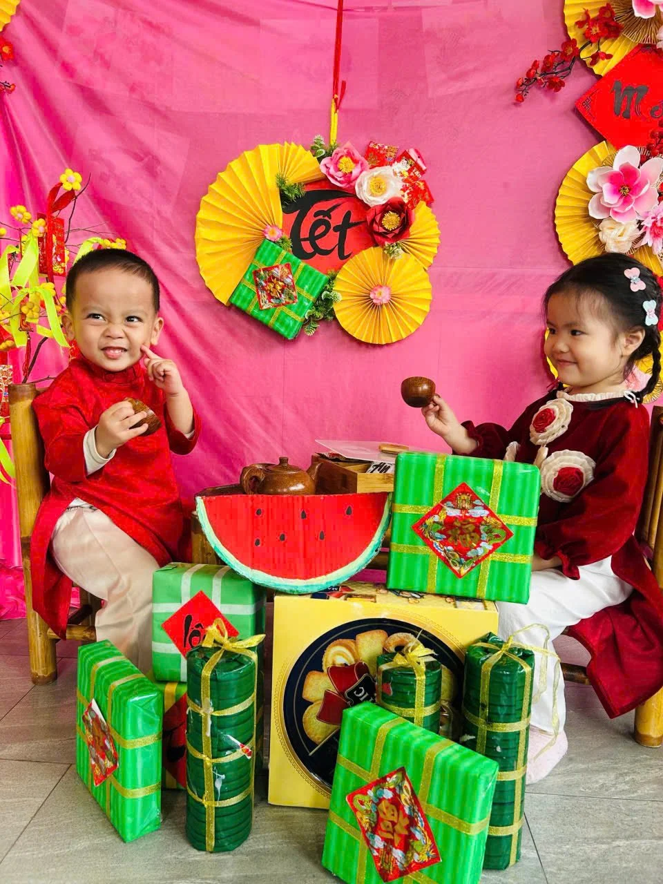 Two children celebrating Lunar New Year, dressed in traditional red clothing, surrounded by gifts wrapped in green paper with red and gold decorations, and holding small bowls. The background features pink and yellow paper fans and decorations.