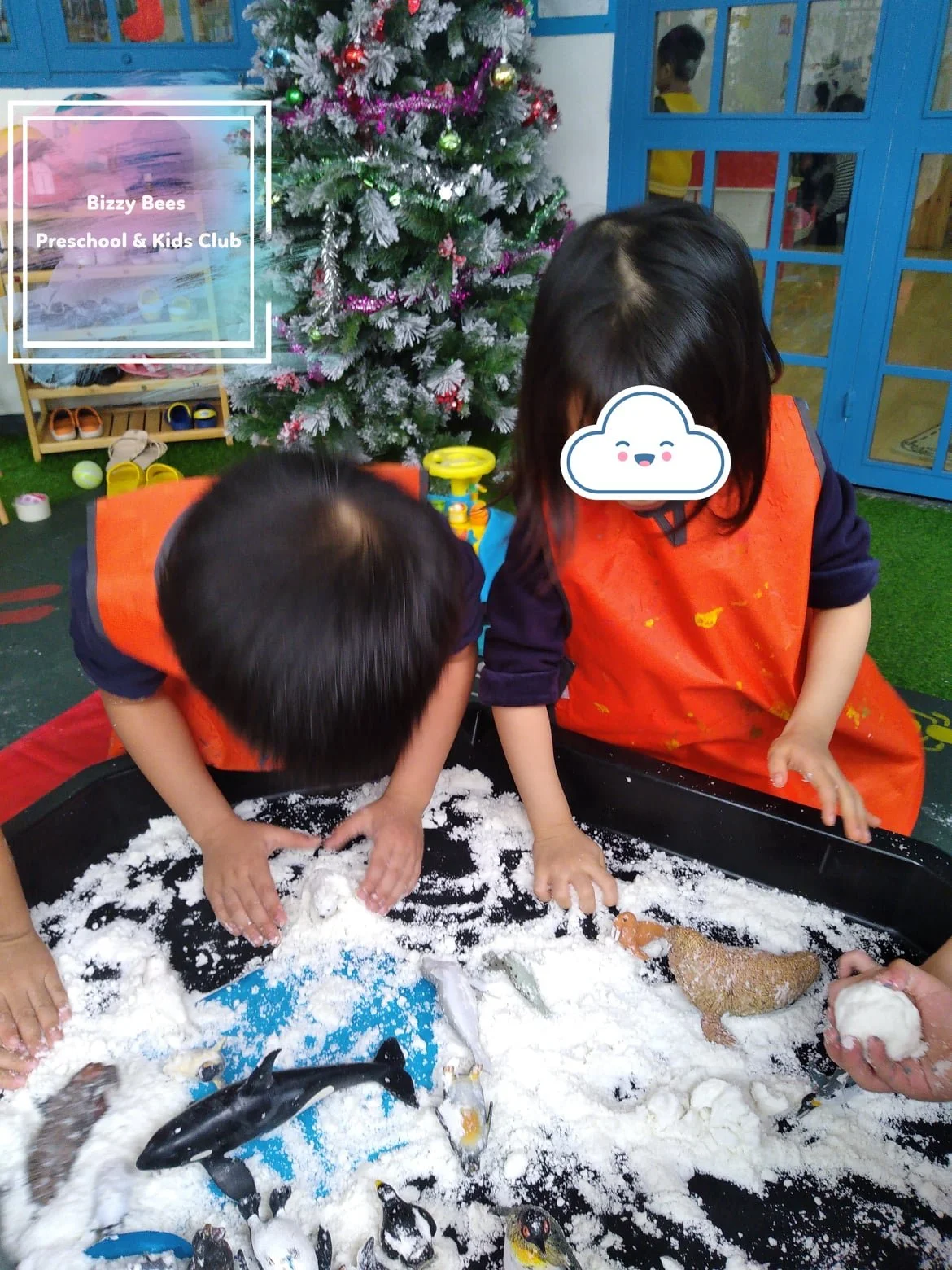 Two young children in orange aprons playing with fake snow and toy penguins in a sensory table, with a decorated Christmas tree in the background at Bizzy Bees Preschool & Kids Club.