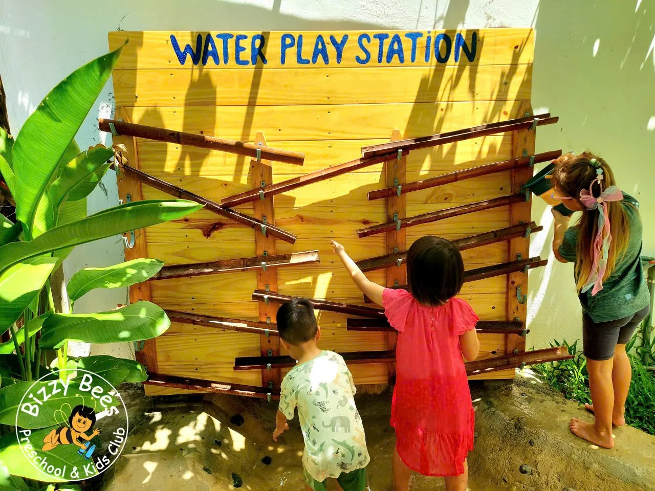 Children playing with a water maze at a water play station, with one child reaching up, another holding a toy camera, and a woman watching, surrounded by green plants.