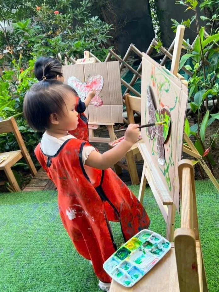 Two young children painting on easels outdoors. One girl is foreground, painting a butterfly with green and purple colors, wearing a red smock. The other girl in the background, also wearing a red smock, is painting a pink and white butterfly. The se
