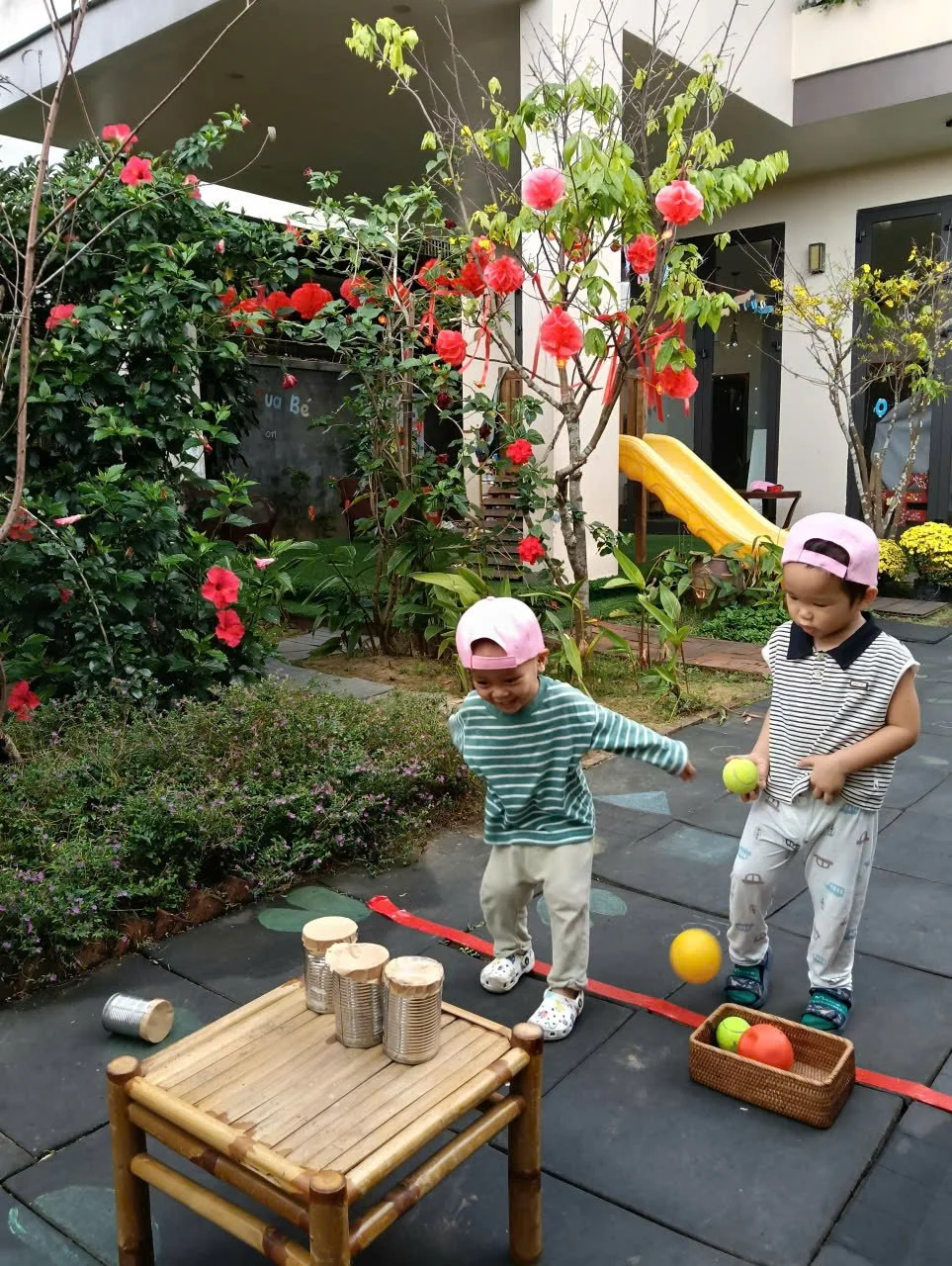 Two young children playing outside in a garden with flowering plants. They are juggling or playing with colorful balls near a small bamboo table and a basket of balls, surrounded by greenery and a small yellow slide.